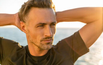 A man with short, styled hair and a beard stands near the ocean, gazing into the distance. He is wearing a black shirt and has his hands placed behind his head. The sun is shining, creating a warm, golden light around him.