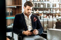 A man with dark hair and a beard is sitting in a stylishly furnished room, looking down at his smartphone. He is dressed in a dark blazer, white shirt, and light gray trousers, with a red pocket square. Shelves filled with various items are in the background.