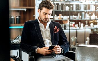 A man with dark hair and a beard is sitting in a stylishly furnished room, looking down at his smartphone. He is dressed in a dark blazer, white shirt, and light gray trousers, with a red pocket square. Shelves filled with various items are in the background.