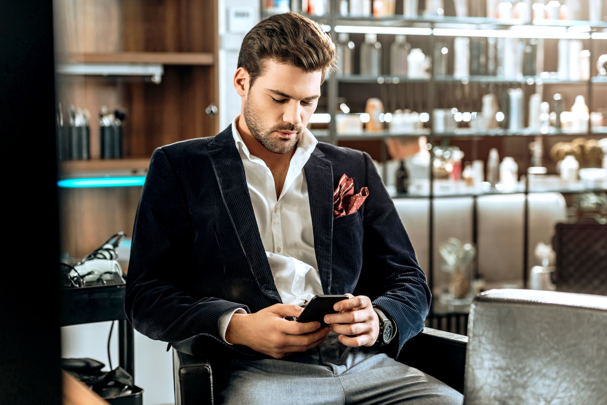 A man with dark hair and a beard is sitting in a stylishly furnished room, looking down at his smartphone. He is dressed in a dark blazer, white shirt, and light gray trousers, with a red pocket square. Shelves filled with various items are in the background.