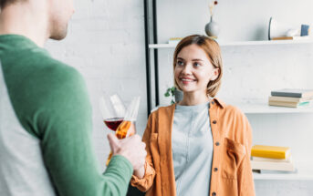 A woman with short hair and wearing an orange shirt smiles while holding a glass of wine and looking at a man with short hair and wearing a green and gray shirt. They are standing in a room with white walls and a bookshelf in the background.