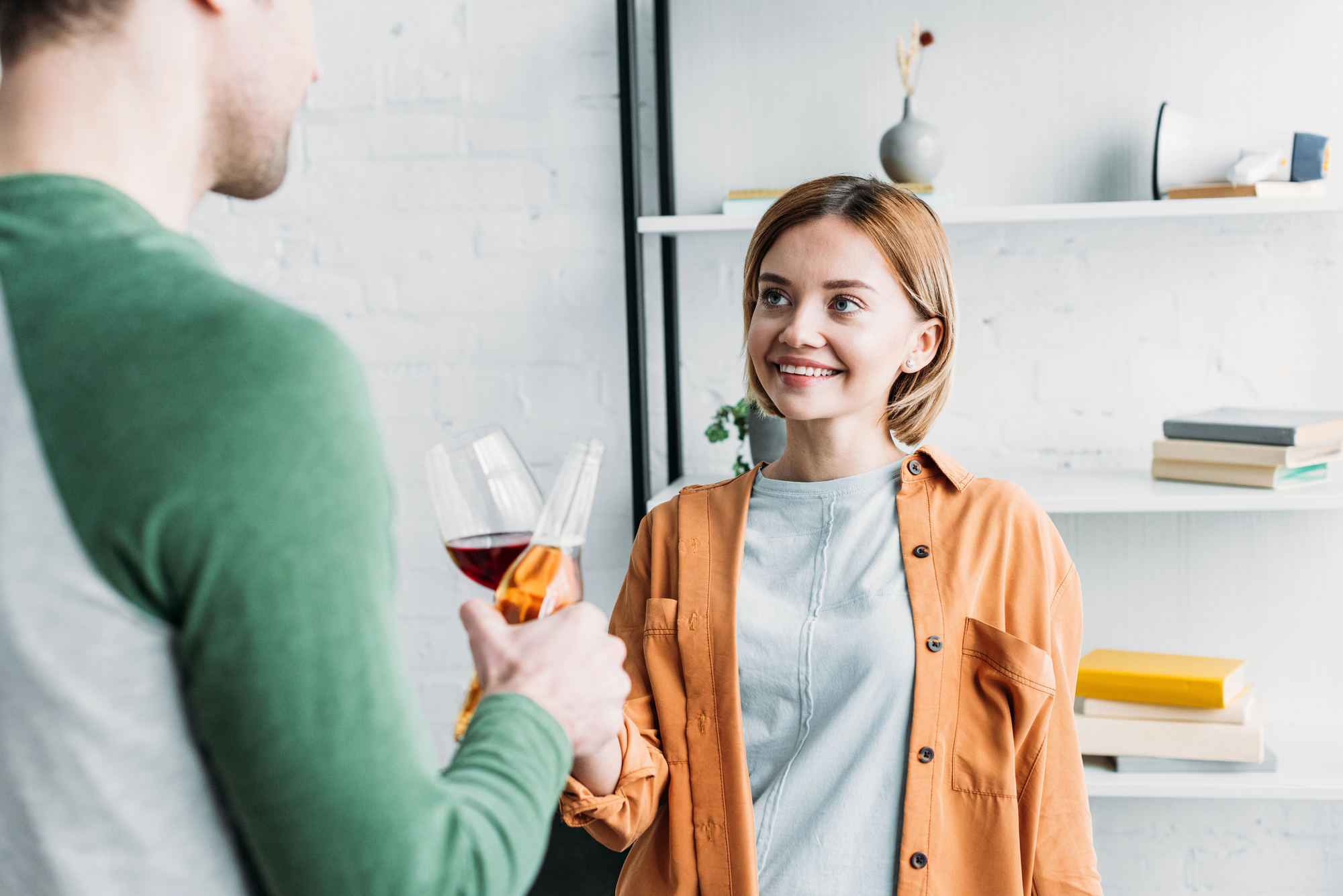 A woman with short hair and wearing an orange shirt smiles while holding a glass of wine and looking at a man with short hair and wearing a green and gray shirt. They are standing in a room with white walls and a bookshelf in the background.