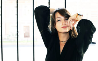 A woman with shoulder-length hair and a slight smile poses with one arm raised and bent at the elbow, resting her hand near her forehead. She's wearing a black top, a necklace, and a watch. The background features vertical bars and a blurred cityscape.