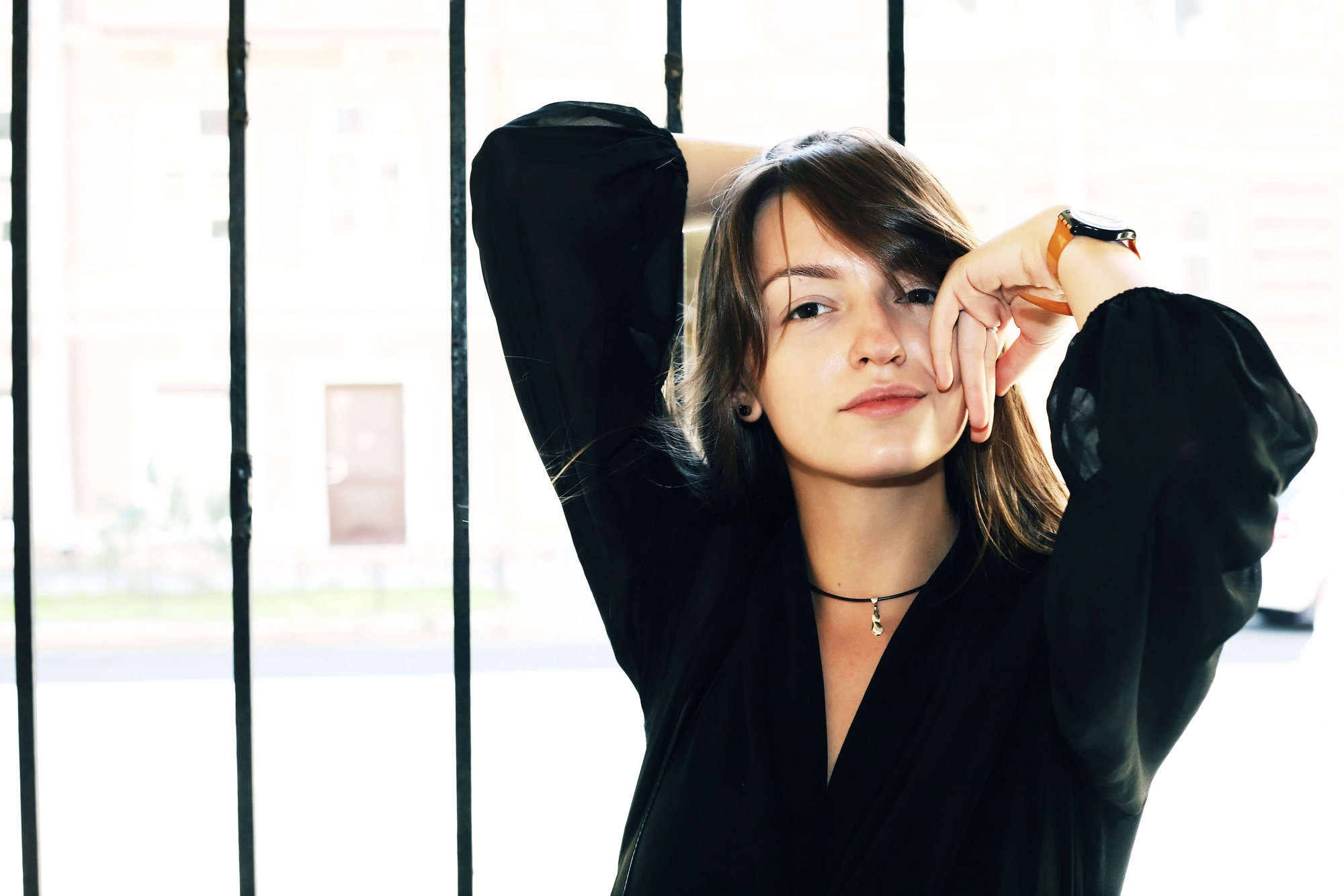 A woman with shoulder-length hair and a slight smile poses with one arm raised and bent at the elbow, resting her hand near her forehead. She's wearing a black top, a necklace, and a watch. The background features vertical bars and a blurred cityscape.