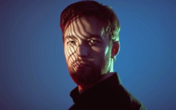 A man with short hair and a beard is illuminated by dramatic, colorful lighting. Shadows cast by a striped pattern partially obscure his face, creating a striking visual effect. He is wearing a dark shirt, and the background is a gradient of blue tones.