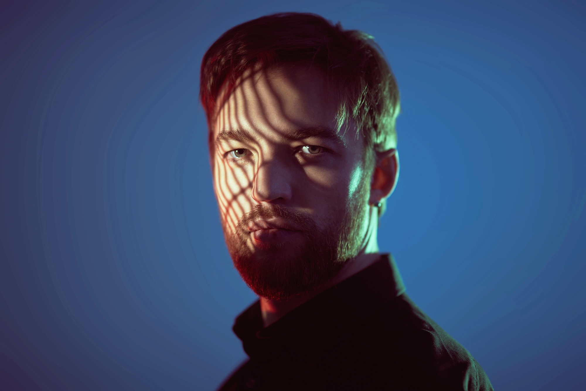 A man with short hair and a beard is illuminated by dramatic, colorful lighting. Shadows cast by a striped pattern partially obscure his face, creating a striking visual effect. He is wearing a dark shirt, and the background is a gradient of blue tones.