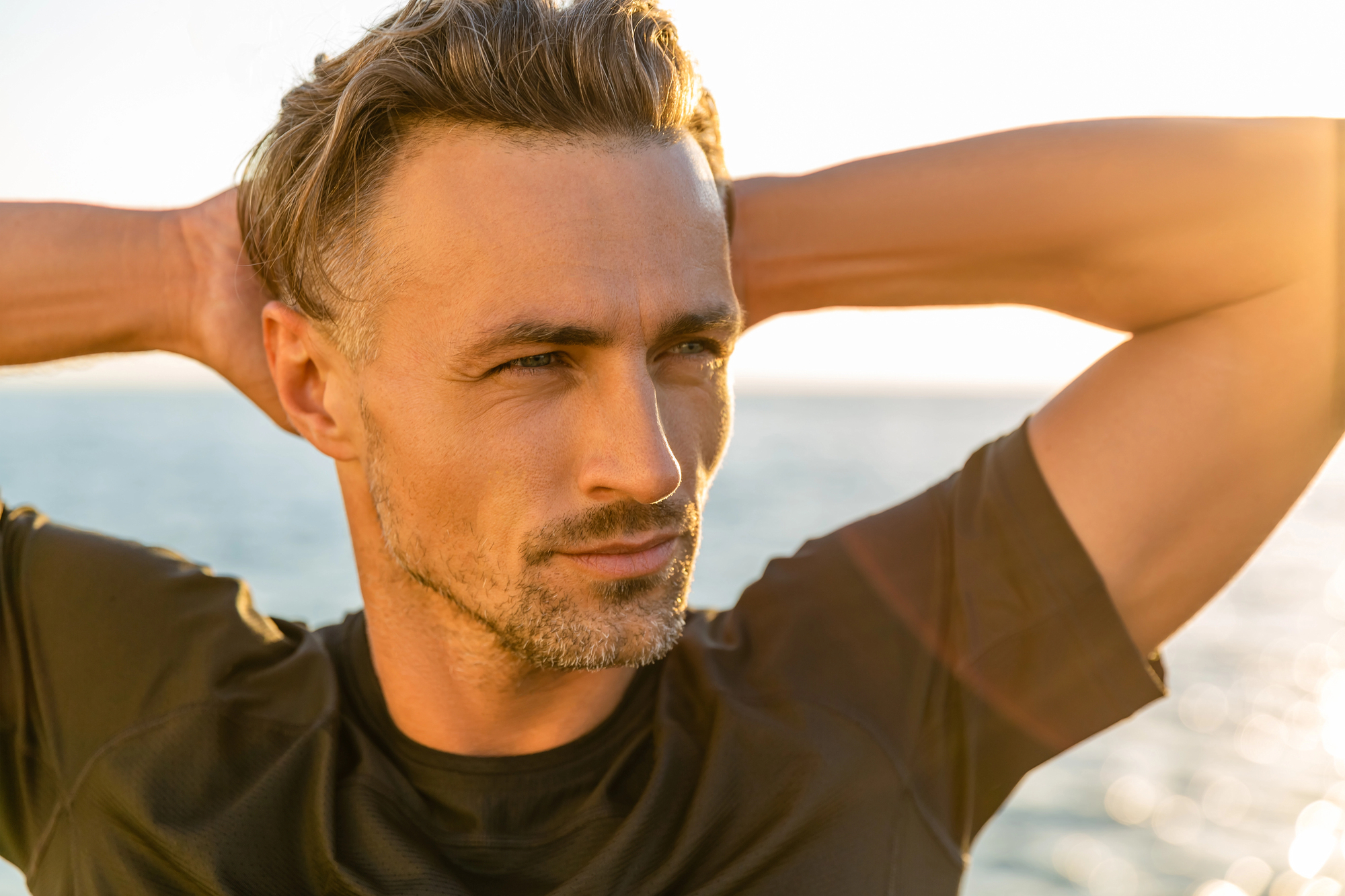 A man with short, styled hair and a beard stands near the ocean, gazing into the distance. He is wearing a black shirt and has his hands placed behind his head. The sun is shining, creating a warm, golden light around him.