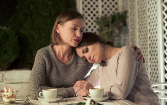 Two women sit at a white outdoor table. One woman, in a gray sweater, embraces the other, who rests her head on the first woman's shoulder. Small desserts and drinks are on the table. The setting appears to be a cozy, lattice-enclosed garden.