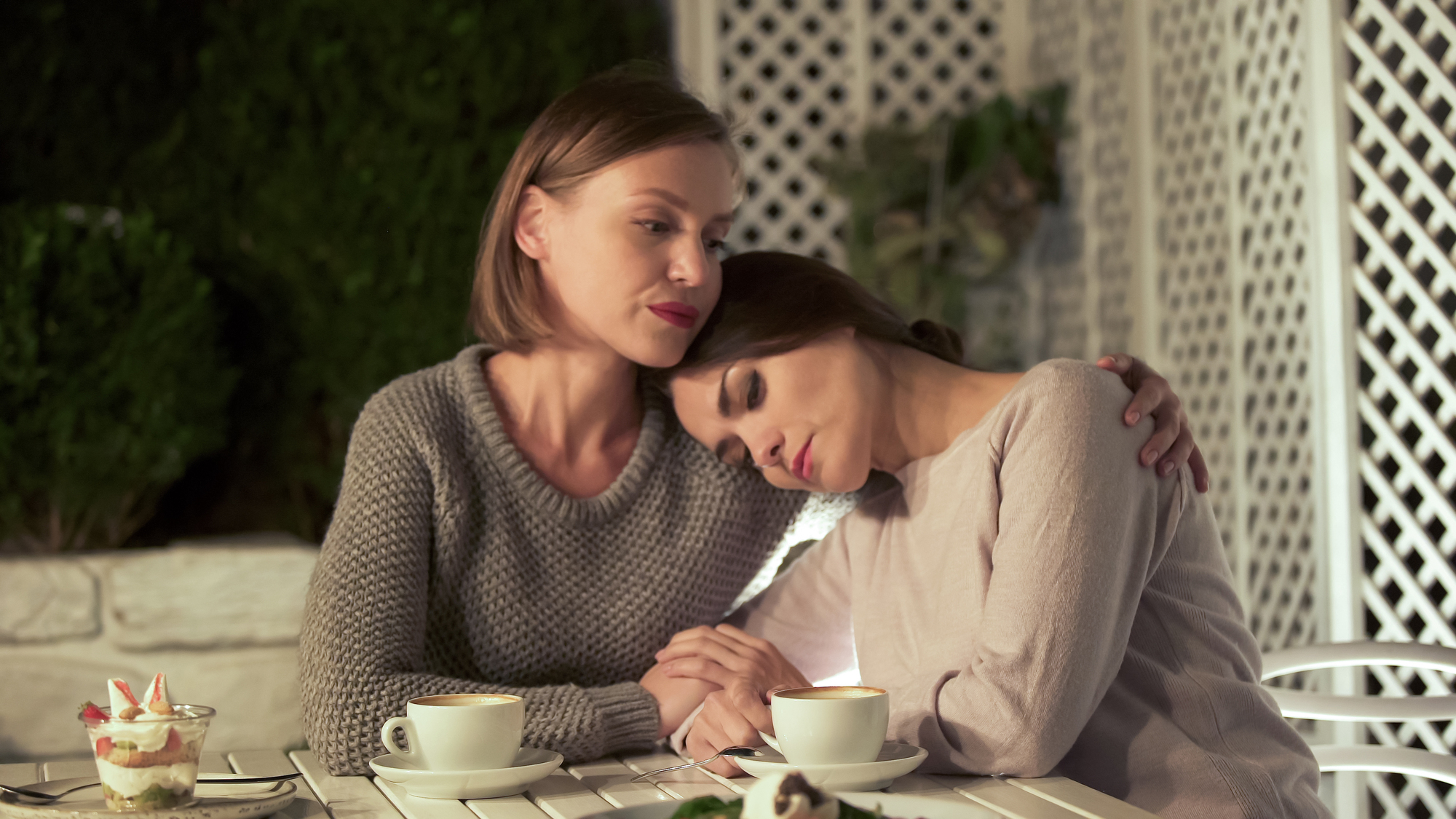 Two women sit at a white outdoor table. One woman, in a gray sweater, embraces the other, who rests her head on the first woman's shoulder. Small desserts and drinks are on the table. The setting appears to be a cozy, lattice-enclosed garden.