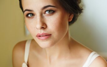 A close-up of a woman with fair skin, green eyes, and dark hair pulled back. She is wearing a light-colored top and has a soft expression, with subtle makeup showcasing her natural freckles and pink lips. The background is softly blurred, drawing focus to her face.