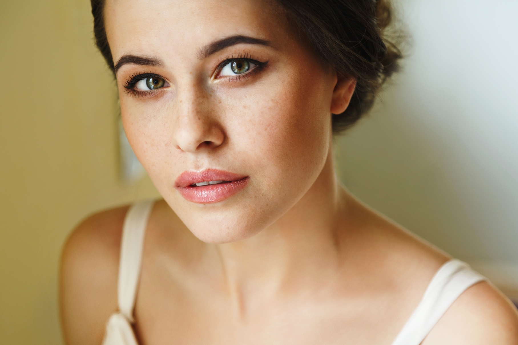 A close-up of a woman with fair skin, green eyes, and dark hair pulled back. She is wearing a light-colored top and has a soft expression, with subtle makeup showcasing her natural freckles and pink lips. The background is softly blurred, drawing focus to her face.