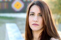 A woman with long, straight brown hair and neutral makeup is looking directly at the camera with a serious expression. She is outdoors with blurred hints of colorful patterns in the background.