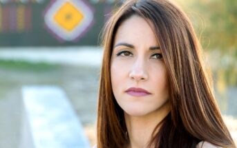 A woman with long, straight brown hair and neutral makeup is looking directly at the camera with a serious expression. She is outdoors with blurred hints of colorful patterns in the background.