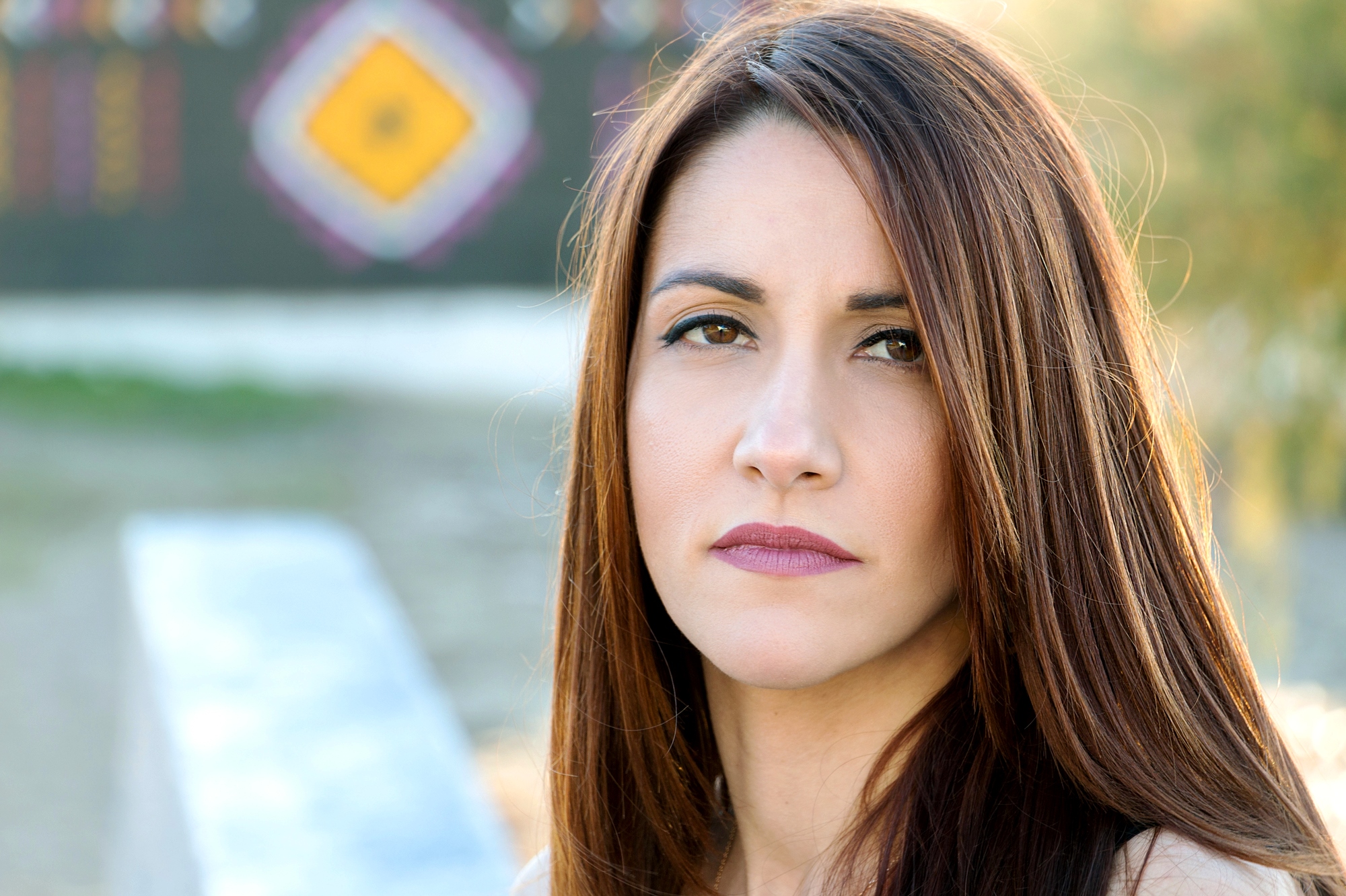 A woman with long, straight brown hair and neutral makeup is looking directly at the camera with a serious expression. She is outdoors with blurred hints of colorful patterns in the background.