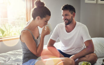 A woman and a man sit closely on a sunlit bed. The woman, in a gray tank top and shorts, sips from a white mug. The man, wearing a white t-shirt and shorts, smiles at her. A bowl of green grapes is placed near them on the bed.