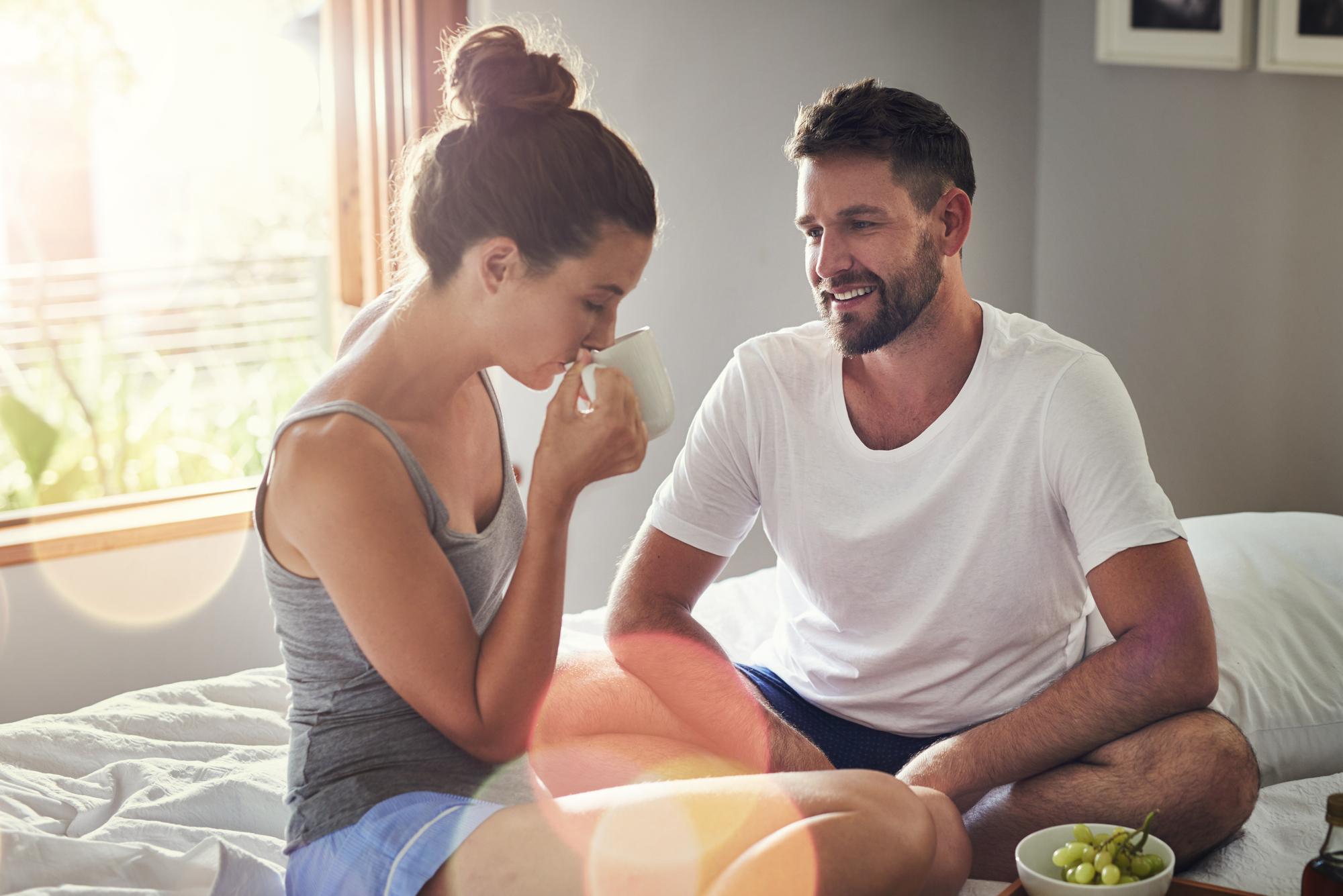 A woman and a man sit closely on a sunlit bed. The woman, in a gray tank top and shorts, sips from a white mug. The man, wearing a white t-shirt and shorts, smiles at her. A bowl of green grapes is placed near them on the bed.