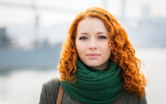 A person with curly, red hair and a green scarf stands outdoors with a blurred urban background. They wear a neutral expression and have a light complexion. The individual is dressed in a grey coat suitable for cool weather.