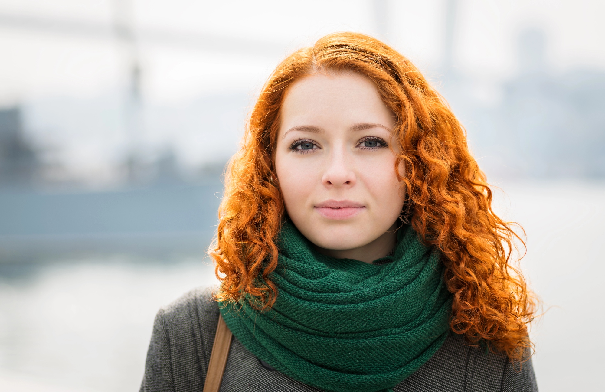 A person with curly, red hair and a green scarf stands outdoors with a blurred urban background. They wear a neutral expression and have a light complexion. The individual is dressed in a grey coat suitable for cool weather.