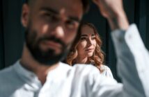 A bearded man with short hair is in the foreground, slightly out of focus, wearing a white shirt. Behind him, a woman with long, wavy hair and a white shirt is in focus, looking sideways and smiling softly. They are in a sunlit room with dark walls.