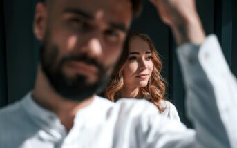 A bearded man with short hair is in the foreground, slightly out of focus, wearing a white shirt. Behind him, a woman with long, wavy hair and a white shirt is in focus, looking sideways and smiling softly. They are in a sunlit room with dark walls.