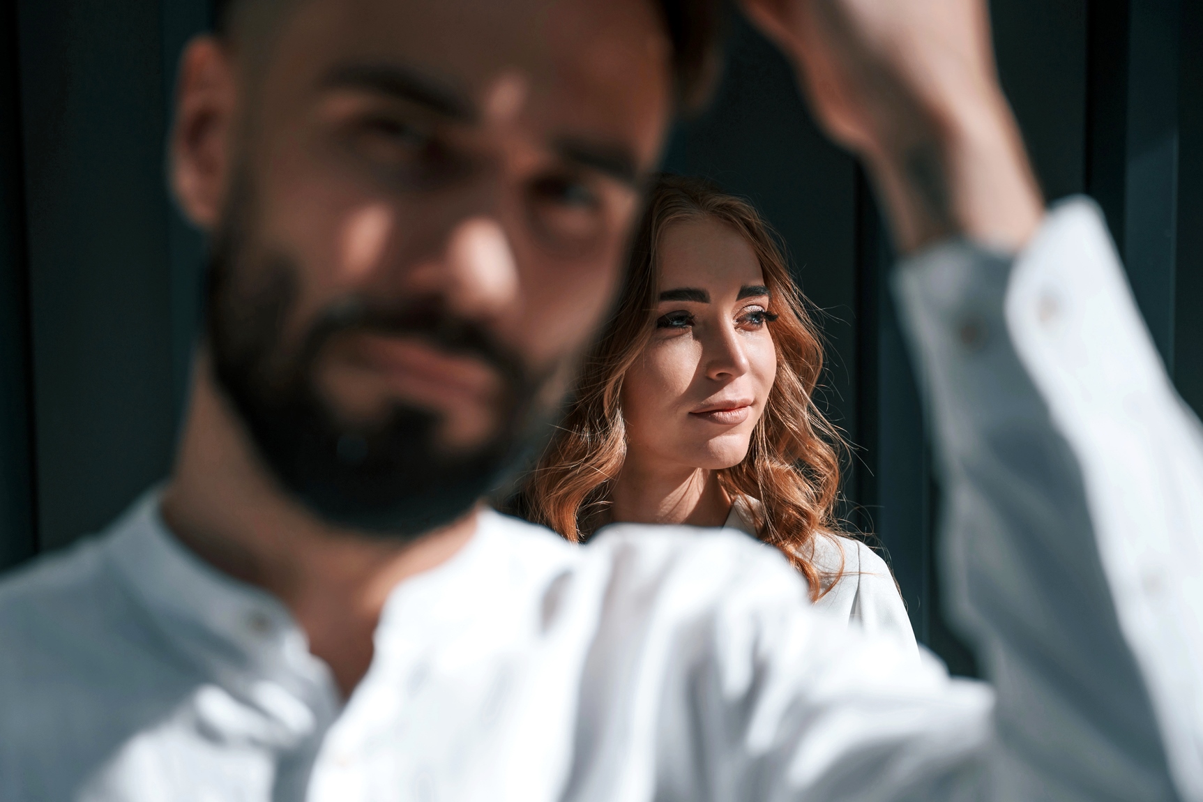 A bearded man with short hair is in the foreground, slightly out of focus, wearing a white shirt. Behind him, a woman with long, wavy hair and a white shirt is in focus, looking sideways and smiling softly. They are in a sunlit room with dark walls.