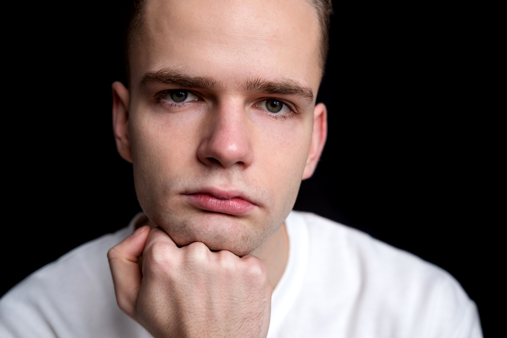 A young man with short hair and a serious expression gazes directly at the camera. He rests his chin on his hand, and is wearing a white shirt. The background is black, creating a strong contrast with his face and clothing.