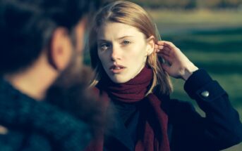 A woman with blonde hair wearing a burgundy scarf and a dark coat stands outdoors, looking towards someone facing her whose back is partially visible. She has her hand behind her ear and a thoughtful expression on her face. The background is slightly blurred.