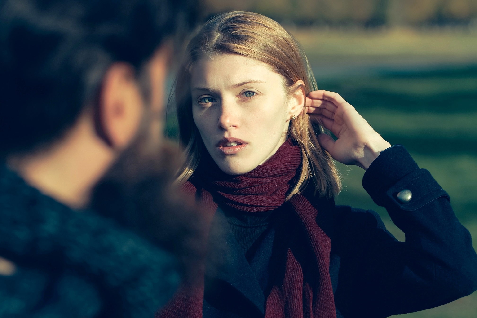 A woman with blonde hair wearing a burgundy scarf and a dark coat stands outdoors, looking towards someone facing her whose back is partially visible. She has her hand behind her ear and a thoughtful expression on her face. The background is slightly blurred.