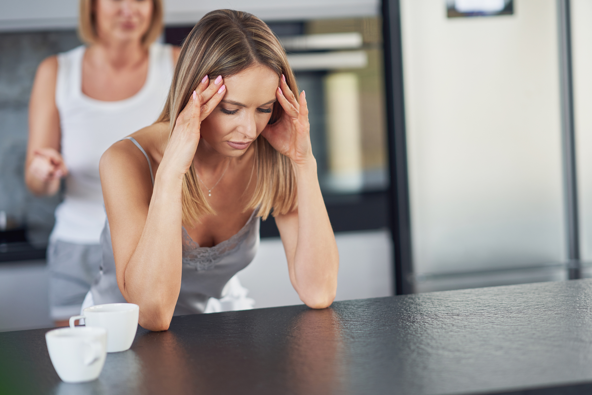 A woman with blonde hair is leaning on a kitchen counter, holding her head with both hands and looking distressed. Another woman in the background appears to be speaking to her. Two white coffee cups are on the counter.