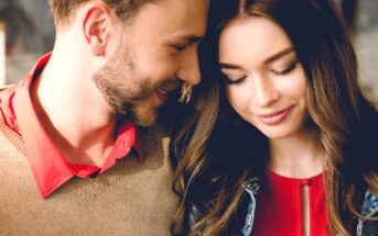 A close-up of a couple, both smiling warmly. The man has a beard and is wearing a brown sweater over a red shirt. The woman has long, wavy hair and is wearing a red top with a denim jacket. They lean close to each other, sharing a tender moment.