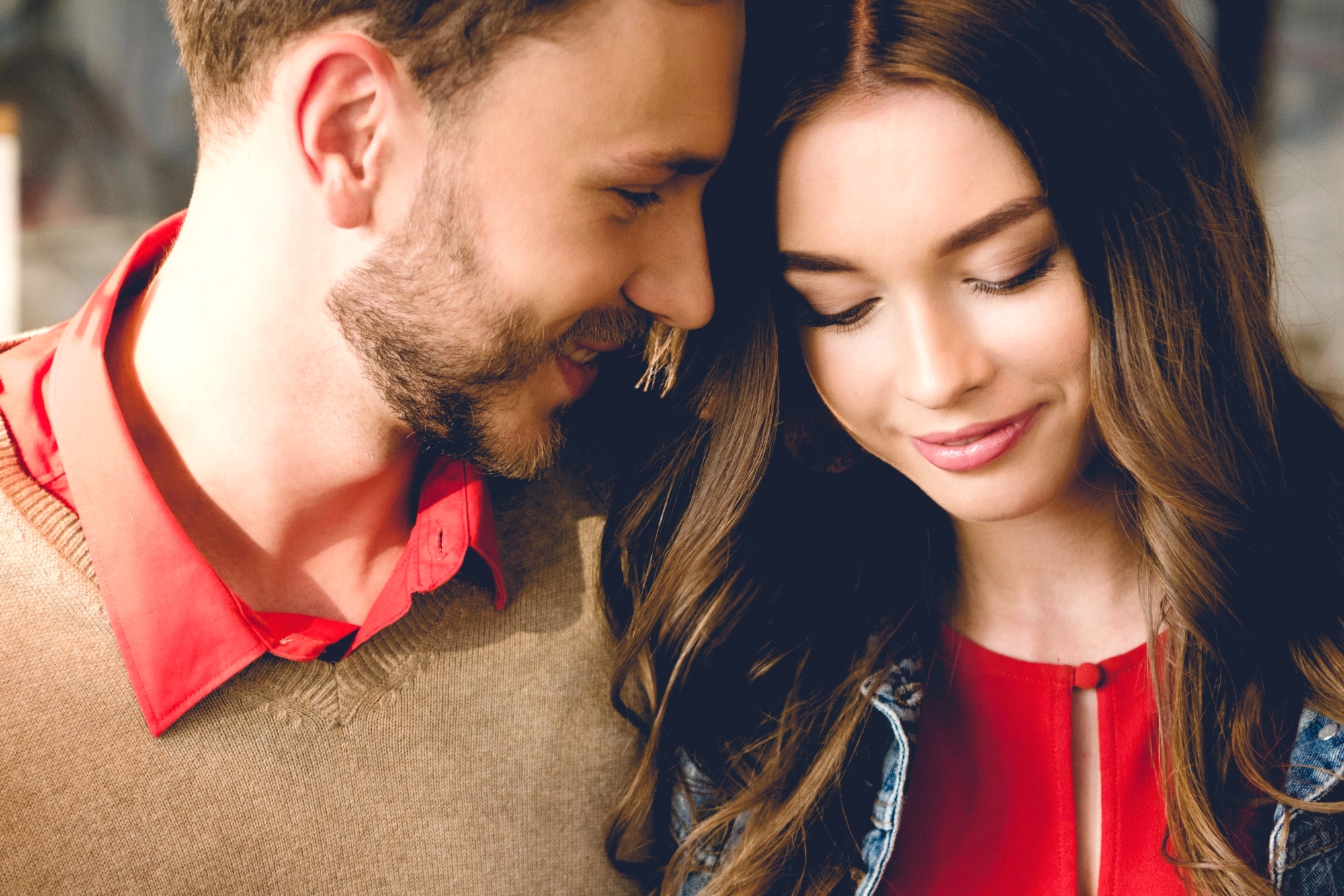 A close-up of a couple, both smiling warmly. The man has a beard and is wearing a brown sweater over a red shirt. The woman has long, wavy hair and is wearing a red top with a denim jacket. They lean close to each other, sharing a tender moment.