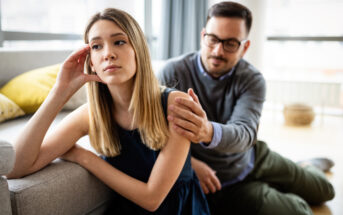 A young woman with long blonde hair sits on the floor, leaning against a sofa, looking upset. A man with glasses and a beard kneels beside her, placing a comforting hand on her shoulder. They appear to be in a bright, modern living room.