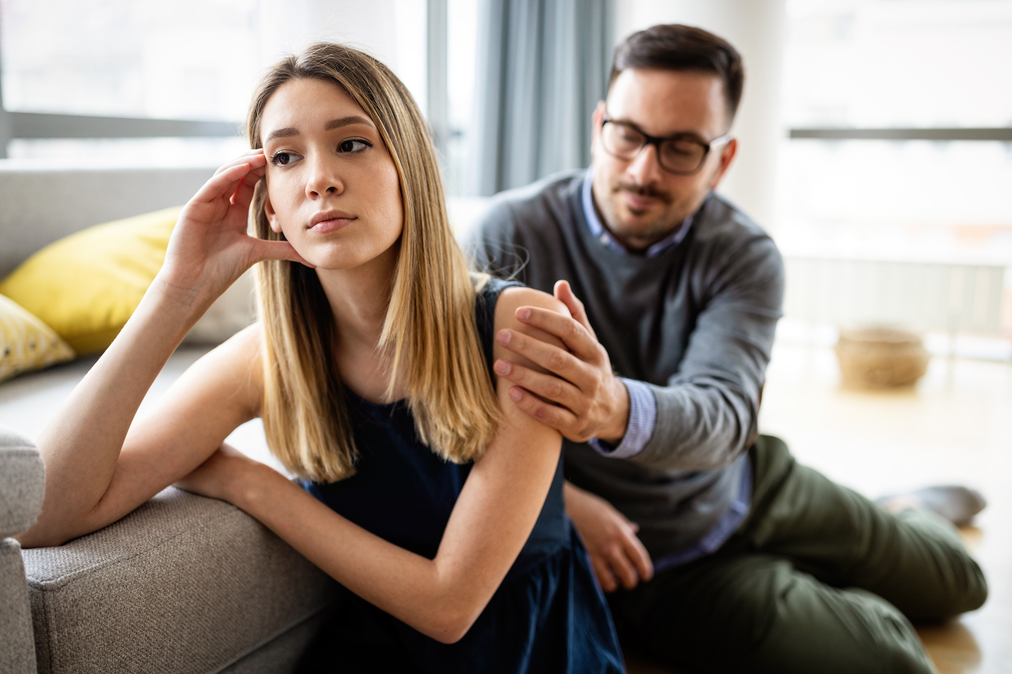 A young woman with long blonde hair sits on the floor, leaning against a sofa, looking upset. A man with glasses and a beard kneels beside her, placing a comforting hand on her shoulder. They appear to be in a bright, modern living room.