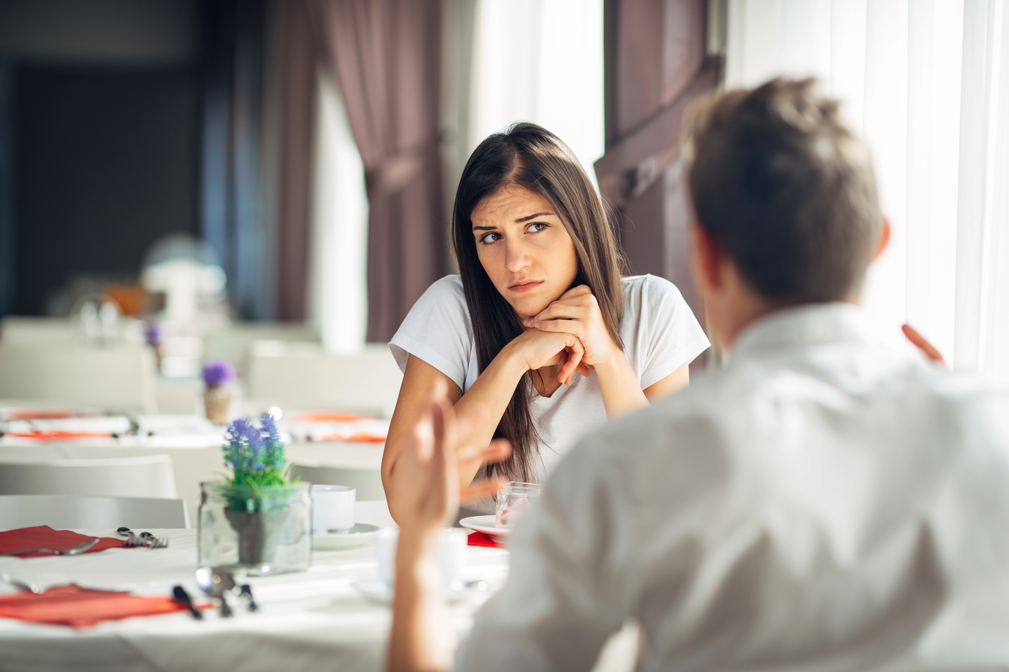 A woman and a man are sitting across from each other at a table in a restaurant. The woman looks concerned, resting her chin on her hands and looking at the man, who is gesturing with his hands. The table has white tablecloths, red napkins, and a small potted plant.