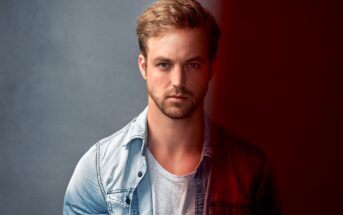 A man with short blonde hair and a beard, wearing a light-colored denim shirt over a gray t-shirt, stands in front of a background that transitions from dark gray on the left to red on the right. He gazes directly at the camera with a neutral expression.