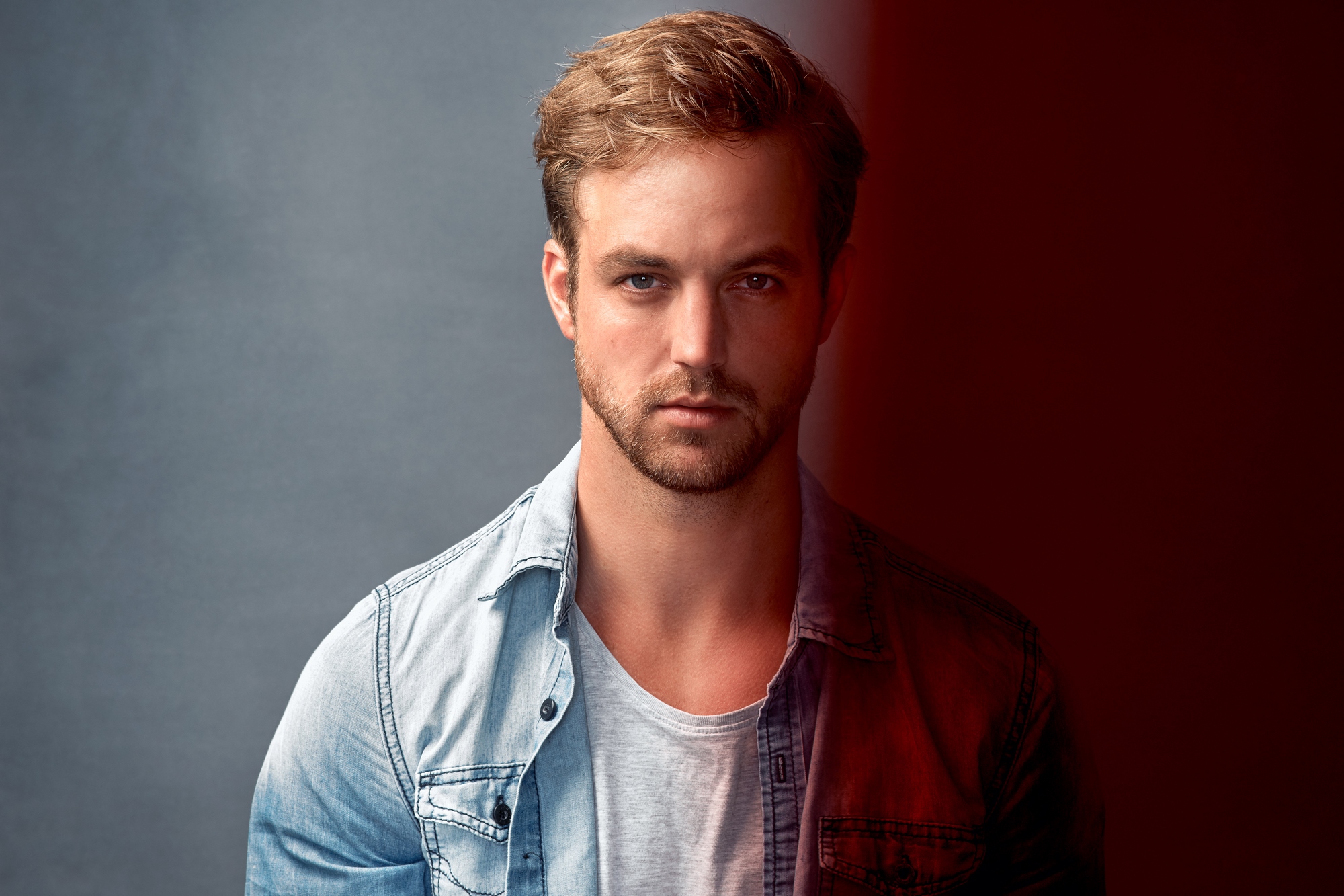 A man with short blonde hair and a beard, wearing a light-colored denim shirt over a gray t-shirt, stands in front of a background that transitions from dark gray on the left to red on the right. He gazes directly at the camera with a neutral expression.