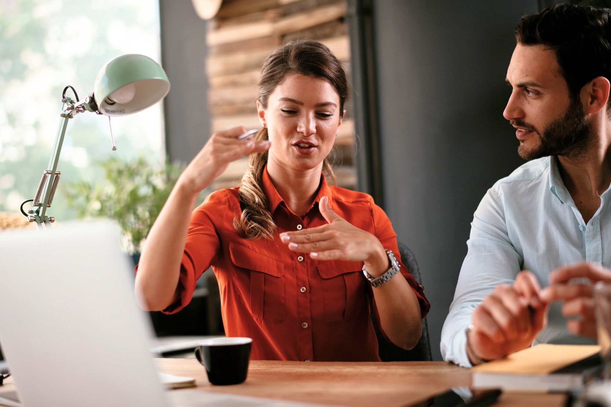 A woman in a red shirt is using sign language to communicate with a man in a light blue shirt at a desk. They are in a modern, well-lit workspace with a laptop, cup, and lamp on the desk. The background includes wooden paneling and a large window.