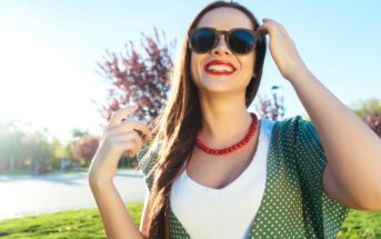 A woman with long brown hair is outdoors, wearing dark sunglasses, a red necklace, a white shirt, and a green polka dot overgarment. She is smiling, with one hand touching her glasses and the other raised near her shoulder. The background has sunlight and trees.