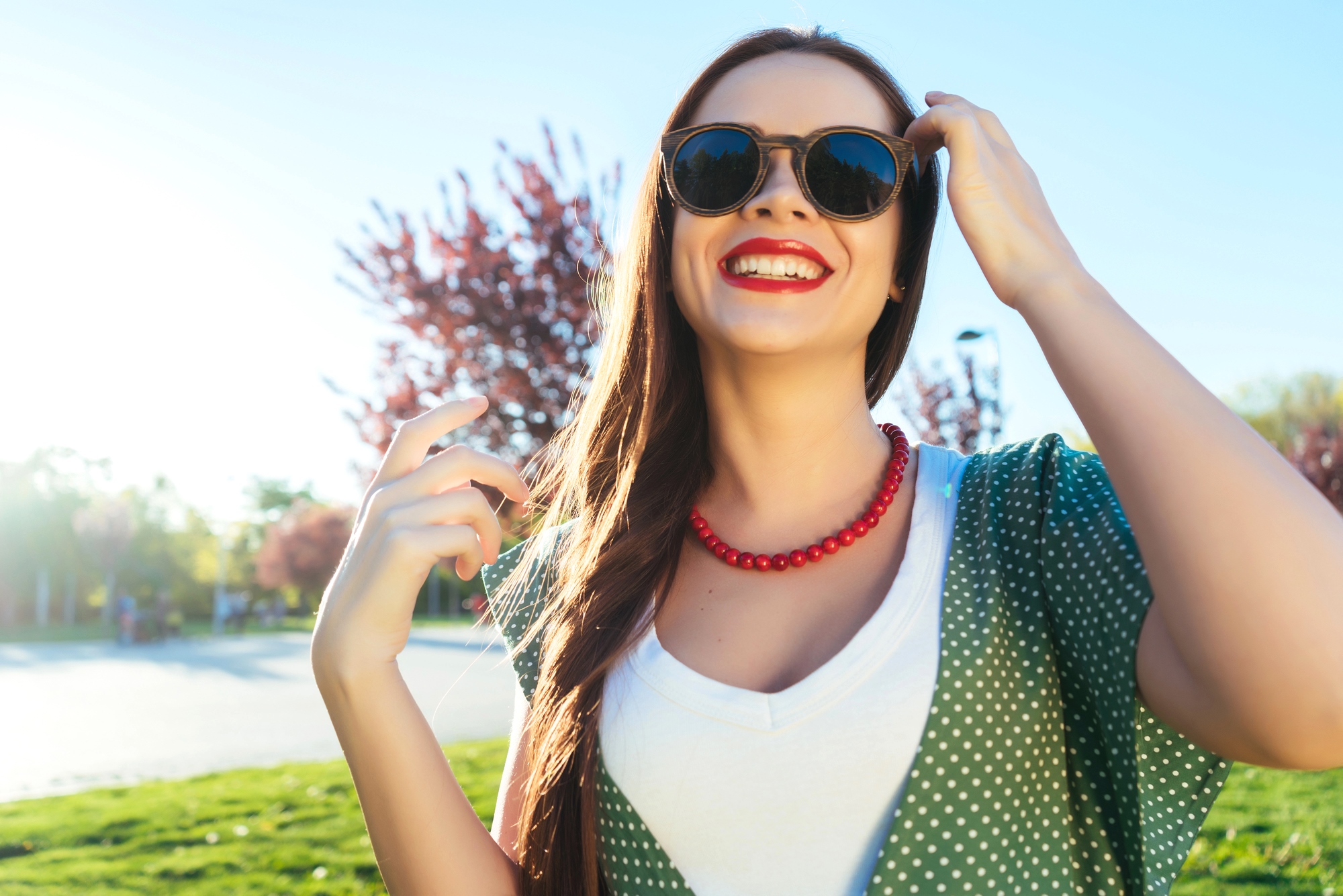 A woman with long brown hair is outdoors, wearing dark sunglasses, a red necklace, a white shirt, and a green polka dot overgarment. She is smiling, with one hand touching her glasses and the other raised near her shoulder. The background has sunlight and trees.