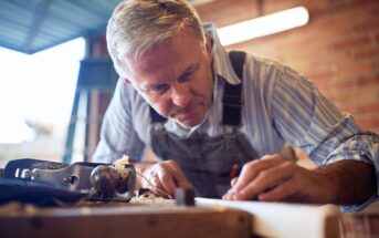 A man with gray hair, wearing a striped shirt and denim overalls, is intensely focused on woodworking. He is using a hand plane on a piece of wood in a workshop setting with a brick wall and a window in the background.