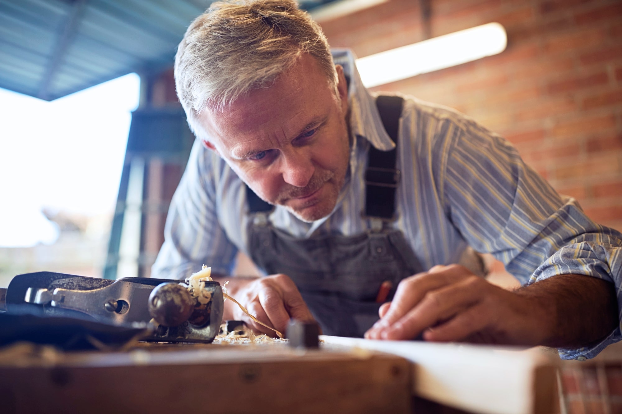 A man with gray hair, wearing a striped shirt and denim overalls, is intensely focused on woodworking. He is using a hand plane on a piece of wood in a workshop setting with a brick wall and a window in the background.