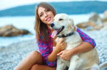 A smiling woman in a pink and blue plaid shirt is sitting on a pebble beach, holding a happy golden retriever. The sea and rocky outcrops are visible in the background.