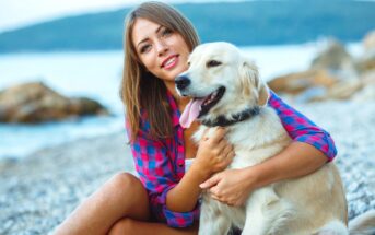 A smiling woman in a pink and blue plaid shirt is sitting on a pebble beach, holding a happy golden retriever. The sea and rocky outcrops are visible in the background.