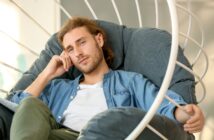 A man with long hair and a beard is sitting in a large, cushioned chair. He wears a blue button-up shirt over a white tee and rests his head on his hand while looking at the camera. The chair has a white, spiral frame. The background is softly lit.