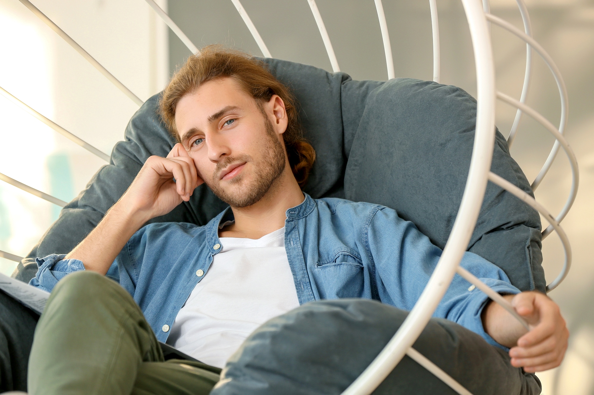 A man with long hair and a beard is sitting in a large, cushioned chair. He wears a blue button-up shirt over a white tee and rests his head on his hand while looking at the camera. The chair has a white, spiral frame. The background is softly lit.