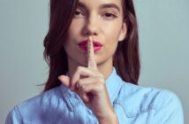 A person with long brown hair wearing a light blue collared shirt holds up their index finger to their lips, signaling for silence. The word "shhh" is written on the finger. The background is plain and gray.