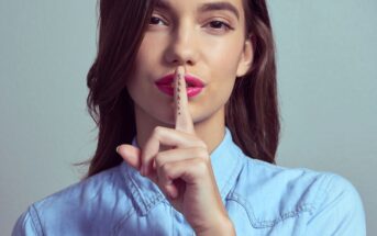 A person with long brown hair wearing a light blue collared shirt holds up their index finger to their lips, signaling for silence. The word "shhh" is written on the finger. The background is plain and gray.