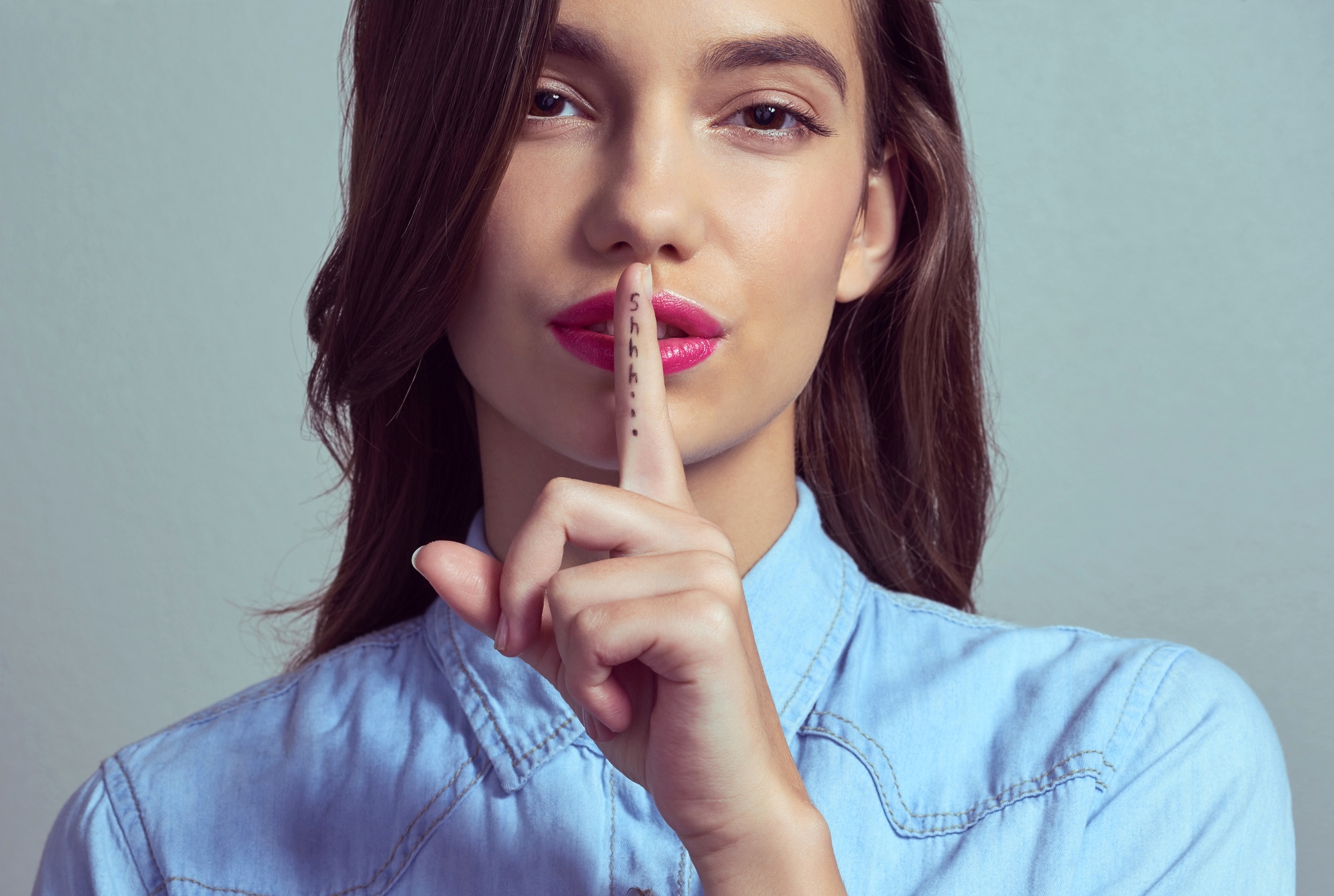 A person with long brown hair wearing a light blue collared shirt holds up their index finger to their lips, signaling for silence. The word "shhh" is written on the finger. The background is plain and gray.