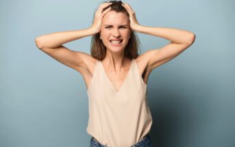 A woman with long hair is standing against a light blue background. She is wearing a beige sleeveless top and blue jeans. Her hands are on her head, her brows are furrowed, and she has a frustrated expression on her face.