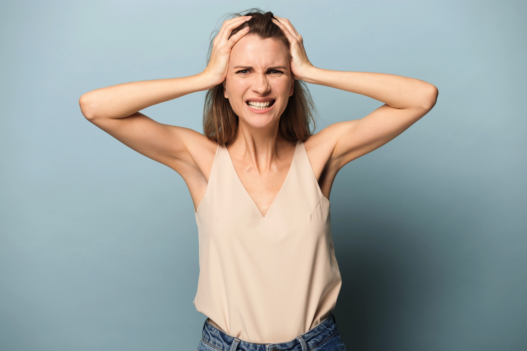 A woman with long hair is standing against a light blue background. She is wearing a beige sleeveless top and blue jeans. Her hands are on her head, her brows are furrowed, and she has a frustrated expression on her face.